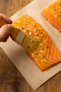 Close-up of brushing garlic butter glaze over fresh salmon fillets on parchment paper.
